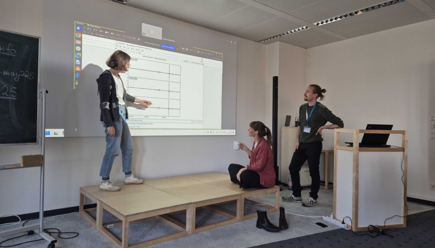Three people in a classroom: one stands on a small wooden stage pointing at a projected diagram, another sits on the stage holding a cup, and a third stands nearby; a laptop on a side stand and a chalkboard frame the scene.