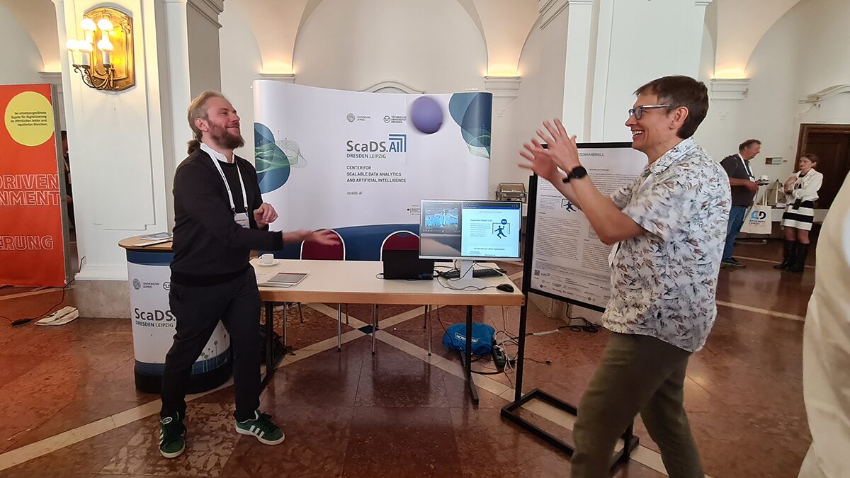 Photo. Thomas Burghard and Robert Haase throwing handball in front of the ScaDS.AI stand, presenting sports analytics in professional handball.