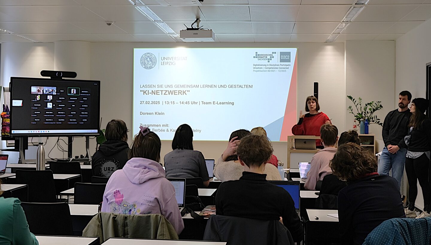 A classroom setting with students seated at desks, facing a projector screen and a large monitor. The projector screen displays a presentation titled "KI-NETZWERK" with details about the event, including the date (27.02.2023), time (13:15 - 14:45 Uhr), and the presenter (Doreen Klein). The large monitor shows a virtual meeting with multiple participants. A person is standing at a podium, speaking, while two others stand nearby. The room has a modern design with white walls, a ceiling-mounted projector, and fluorescent lighting.