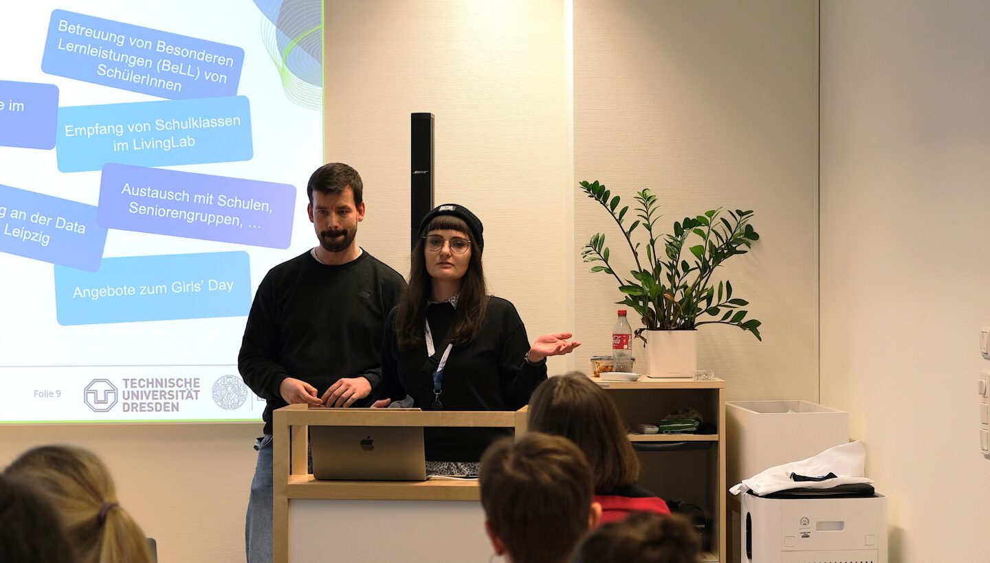 Two people are standing behind a podium in a classroom setting, giving a presentation. The faces of the individuals are obscured for privacy. Behind them, a projection screen displays various text boxes in German, listing different activities and programs. The logo of Technische Universität Dresden is visible at the bottom of the screen. There is a plant and a bottle of soda on a shelf to the right of the podium. Several students are seated in the foreground, facing the presenters.