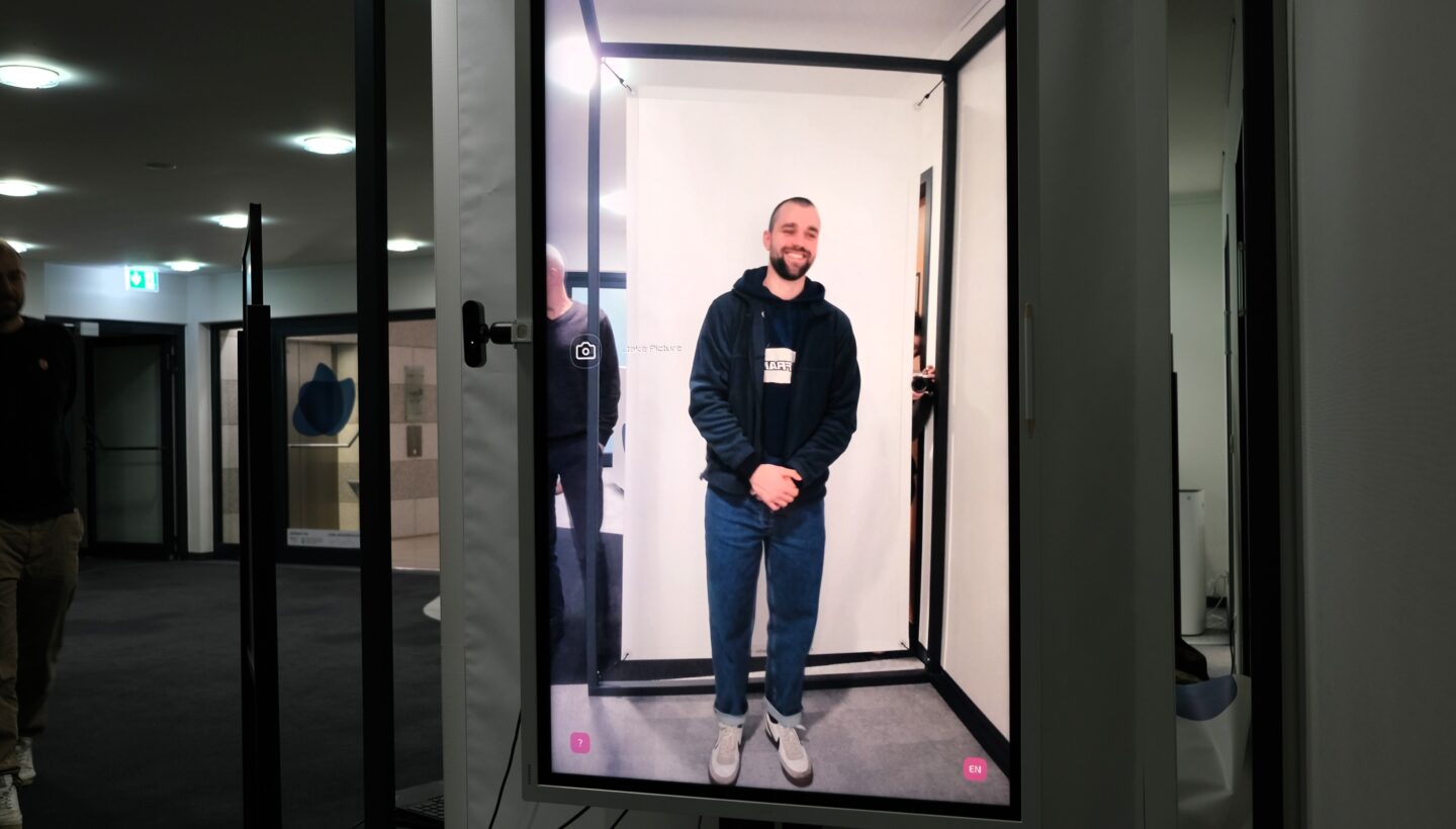 A screen displaying a person standing inside a rectangular frame. The camera is positioned on the left side of the screen. In the background, there are multiple ceiling lights and doors, indicating an indoor setting. The person inside the frame is wearing a dark hoodie, blue jeans, and white sneakers.