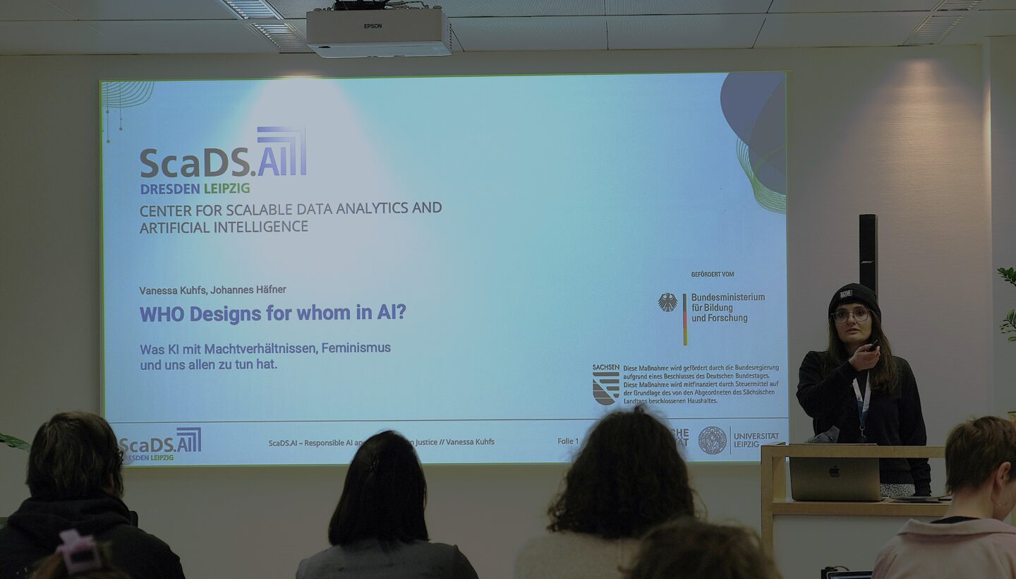 The image shows a presentation slide projected on a screen in a conference room. The slide is titled "WHO Designs for whom in AI?" and is presented by Vanessa Kuhfs and Johannes Häfner. The subtitle reads, "Was KI mit Machtverhältnissen, Feminismus und uns allen zu tun hat," which translates to "What AI has to do with power relations, feminism, and all of us." The presentation is part of an event organized by ScaDS.AI Dresden/Leipzig, the Center for Scalable Data Analytics and Artificial Intelligence. The slide also includes logos of the Bundesministerium für Bildung und Forschung (Federal Ministry of Education and Research), the University of Leipzig, and other supporting organizations. The presenter is standing at a podium on the right side of the image, holding a microphone. Several audience members are visible from behind, seated and facing the screen.