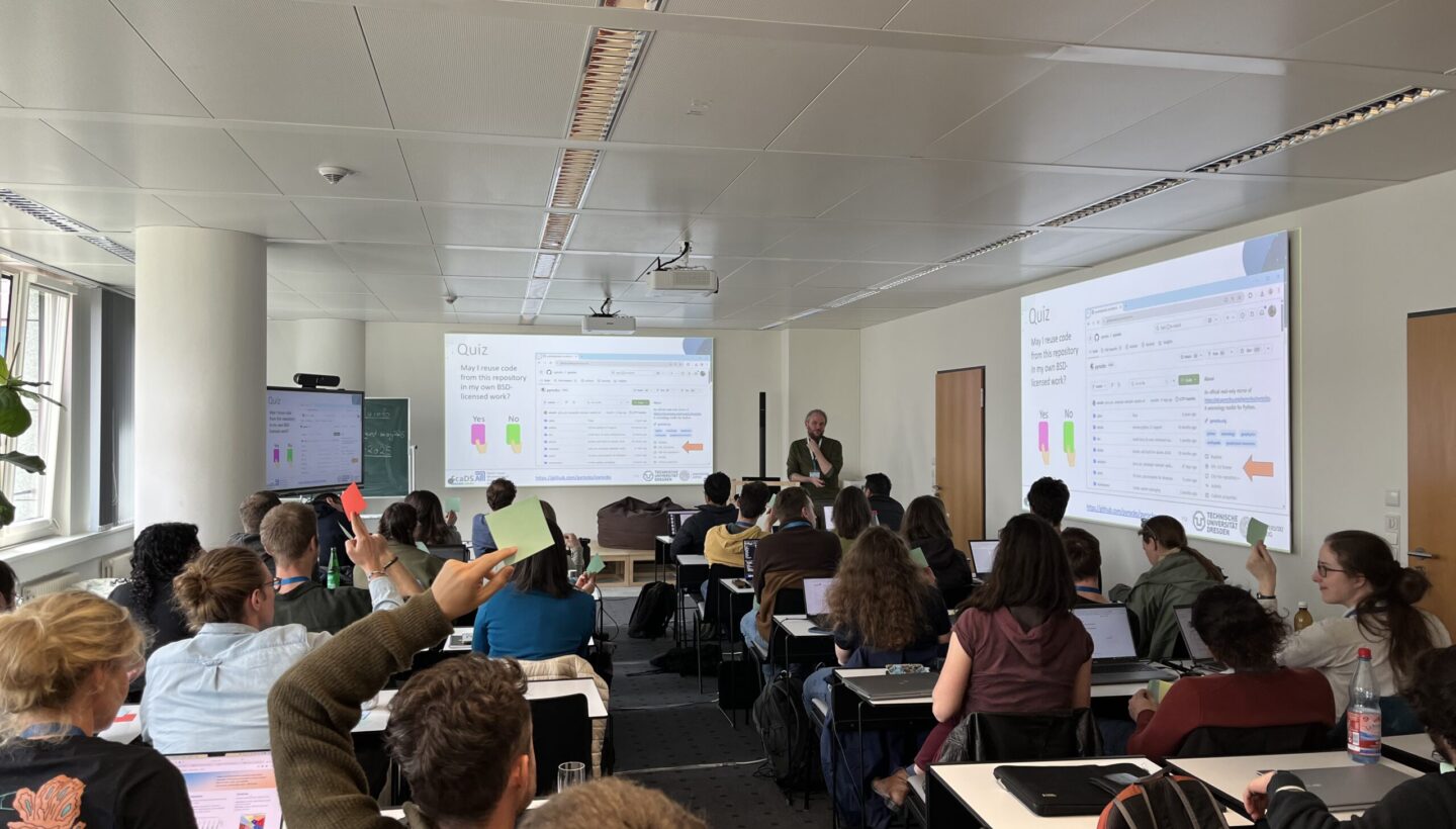 The classroom in the Living Lab Leipzig. People sitting on small tables, raising their hands with either red or green colored papers. All the way in the back is Robert Haase, holding the License Quiz. On the right and back wall is the presentation projected.