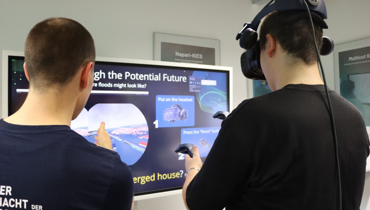 Photo. One person is wearing a VR headset and holding controllers, while another person stands beside them, pointing at a large screen. The screen shows instructions for the VR experience of FloodVis.