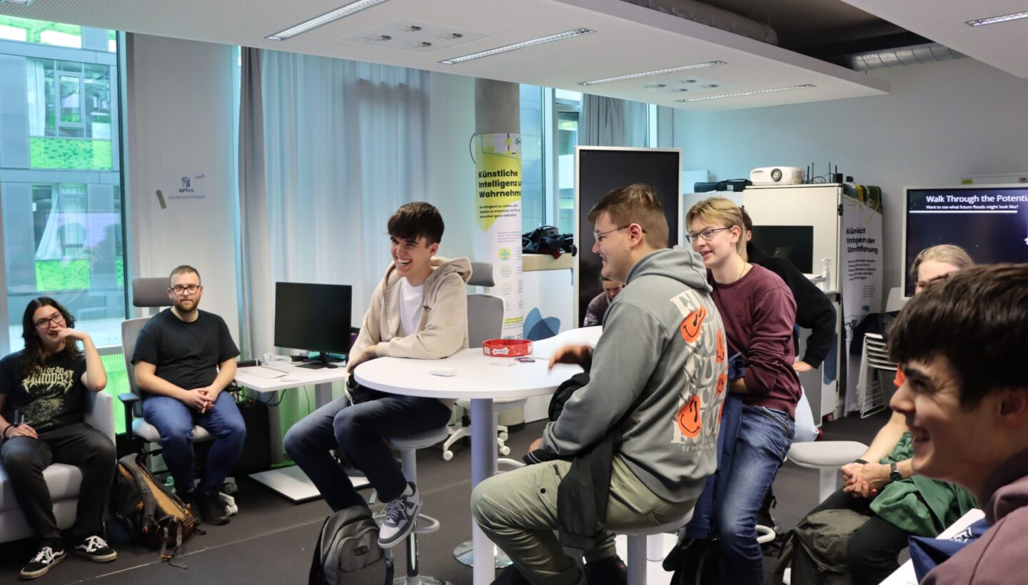 Photo. A group of people are gathered in Living Lab. Two people sitting at a central white table are smiling and laughing, while others relax on a nearby couch and chairs, creating a casual, social atmosphere.