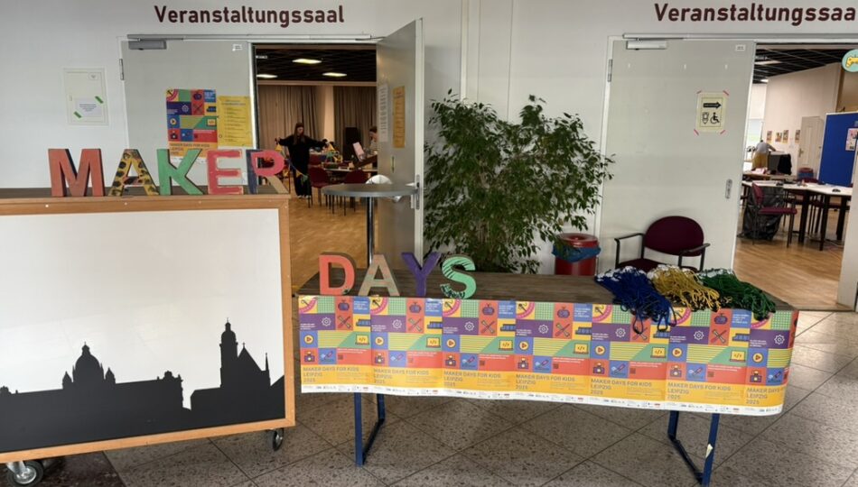 Entrance to an event hall with double doors labeled "Veranstaltungssaal"; a decorated display table in front shows large colorful letters spelling "MAKER DAYS", piles of blue, green, and yellow lanyards, sponsor logos on a banner, a cityscape silhouette board to the left, and a potted plant between the doors.