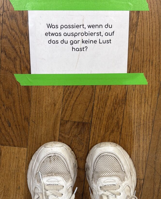 A pair of white sneakers on a wooden floor, positioned in front of a sheet of paper taped down with green tape. The paper displays the German question “Was passiert, wenn du etwas ausprobierst, auf das du gar keine Lust hast?” (“What happens when you try something that you really don’t feel like doing?”).