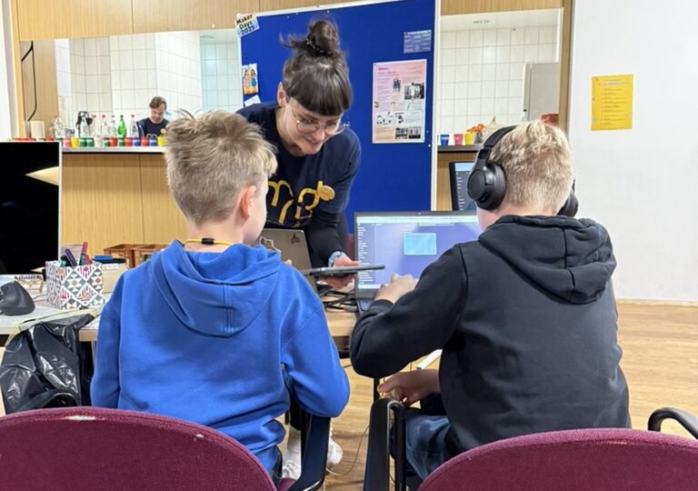 Two children from behind sitting at a table on red velvet chairs and working at laptops. The child on the right side is wearing headphones. A volunteering person wearing a Maker Days for Kids shirt is leaning over the table, with a tablet in hand. In the background is a blue partition and behind that a broad hatch into a kitchen. Aligned on the countertop of the hatch are several colorful cups and water bottles.
