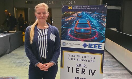 Miriam Louise Carnot stands next to a poster presenting the 37th IEEE Intelligent Vehicles Symposium in Detroit.