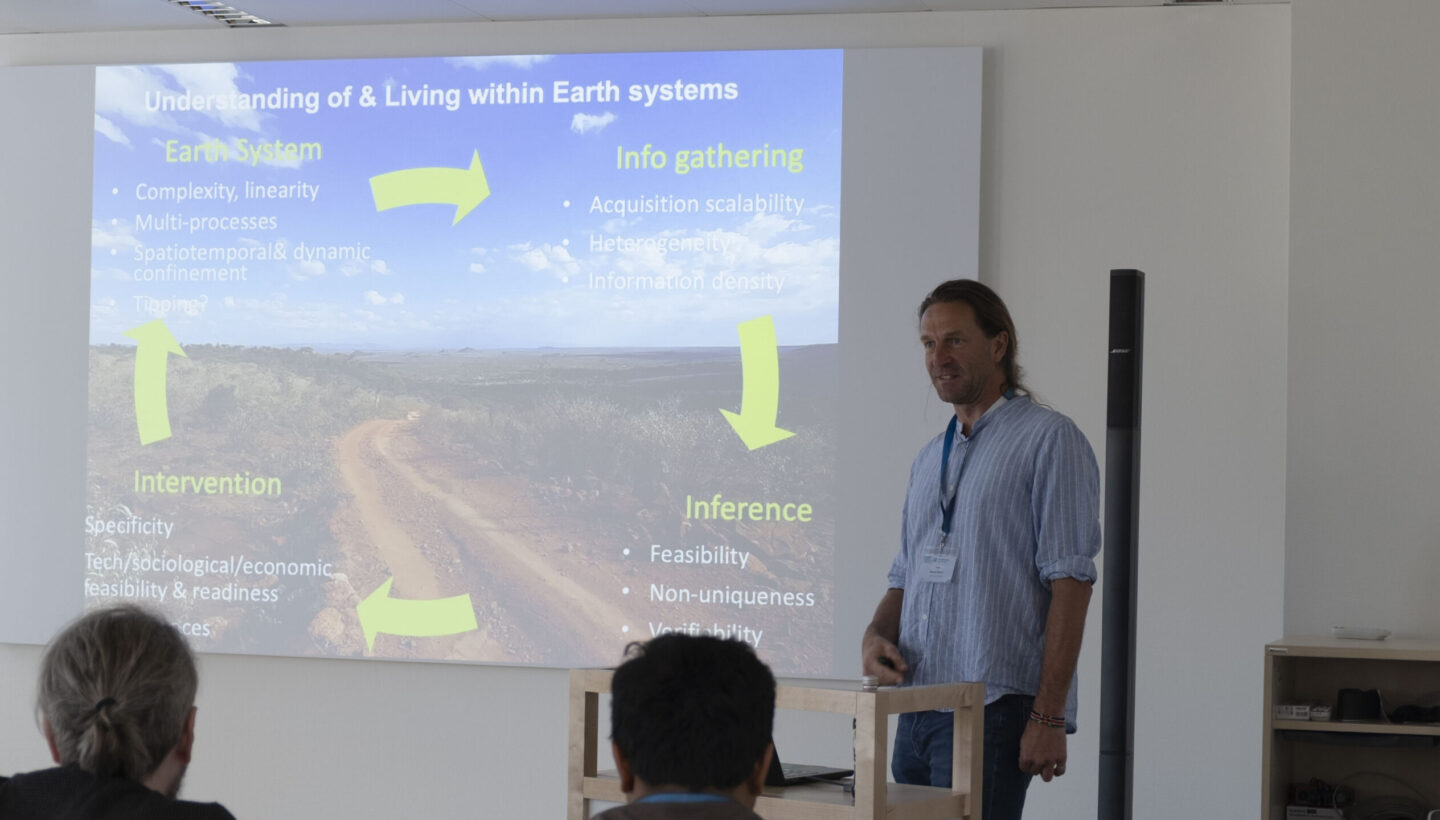 A speaker presents a slide titled "Understanding & Living within Earth Systems." The slide divides the concept into four parts—Earth System, Info Gathering, Inference, and Intervention—connected by arrows, with a natural landscape featuring a dirt road in the background.