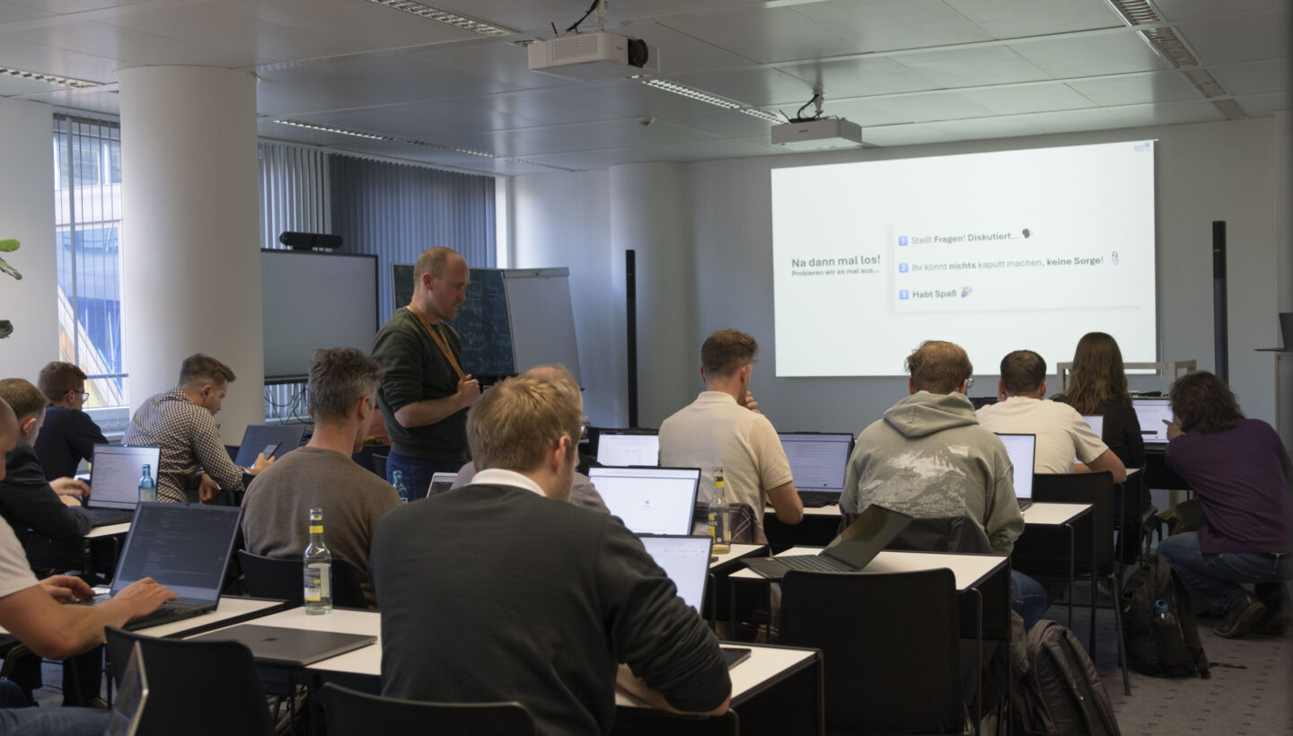 Workshop at the AI Agents & Agent Frameworks Meetup: A dynamic classroom setting where technology enthusiasts use laptops and collaborate near a whiteboard. A projector displays German instructions, highlighting an interactive session on AI agents and intelligent automation.