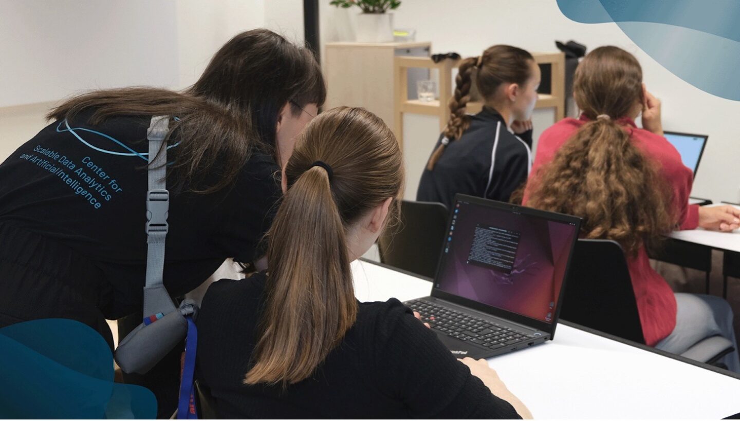 Photo. A ScaDS.AI employee leans over the shoulder of a pupil and helps her with her work on a laptop at a workshop to support with the preparation of the Komplexe Leistung. Other students can be seen in the background, also working on laptops. The room looks like a modern classroom where practical exercises on AI technologies take place.