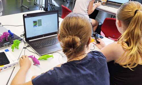 Photo. Two schoolgirls are sitting at a table in front of a laptop with an AI application open. Next to the laptop are colourful, 3D-printed figures.