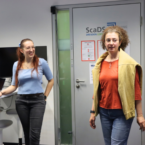 A woman in an orange top with a beige sweater over her shoulders stands beside another woman in a blue top during TUD Freshers’ Week 2026 at the ScaDS.AI Dresden/Leipzig Living Lab.
