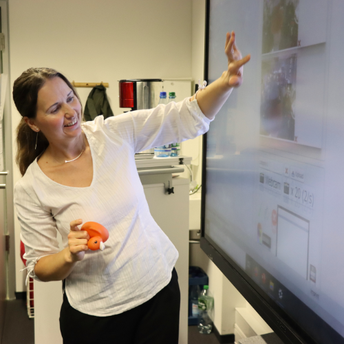 A woman in a white blouse smiling and gesturing toward an interactive touchscreen during TUD Freshers’ Week 2026 at the ScaDS.AI Dresden/Leipzig Living Lab.