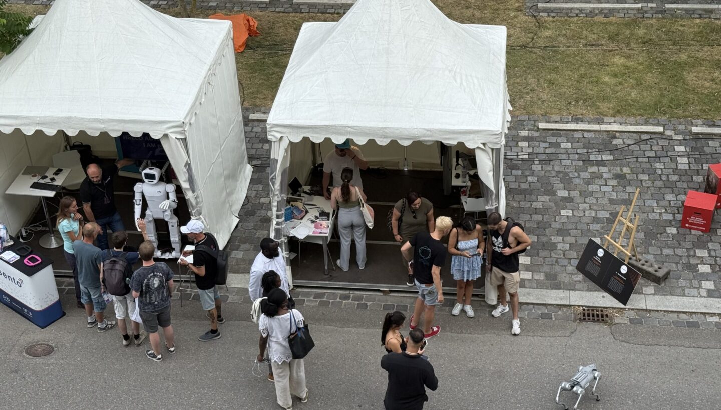 Two white canopy tents stand on a paved outdoor plaza at a robotics exhibition. Under the left tent, a humanoid robot is positioned beside a table of equipment while several people observe and interact. The right tent contains another display table where visitors converse and explore items. Nearby on the ground, a small four-legged robot waits as more attendees mill around the area.