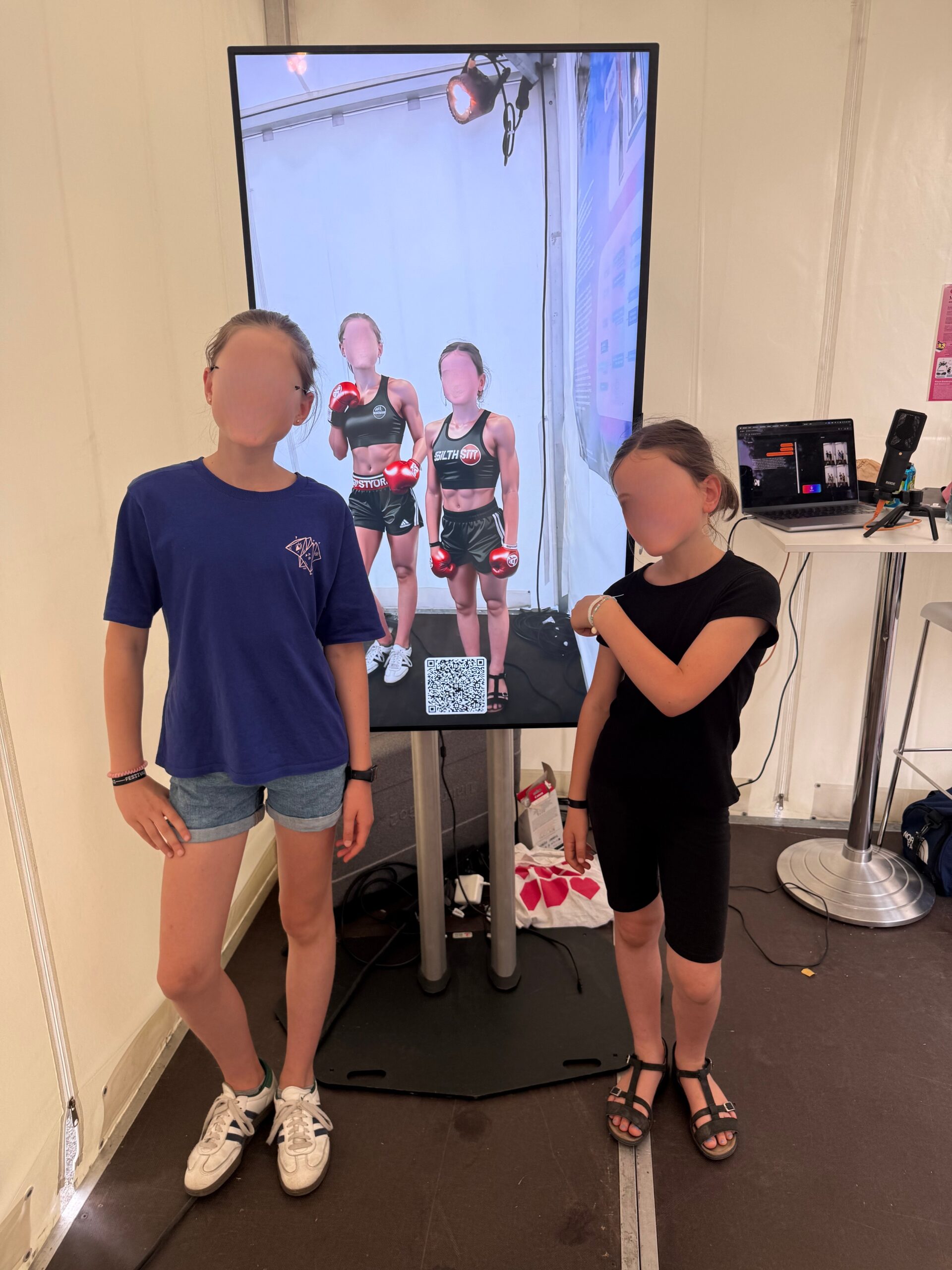 Two children stand side by side facing a tall vertical touchscreen display - the magic mirror - that shows a photo of the two children in boxing gloves and shorts, with a QR code along the bottom. To the right of the screen is a small table holding a laptop and other equipment, all set against a light-colored backdrop.