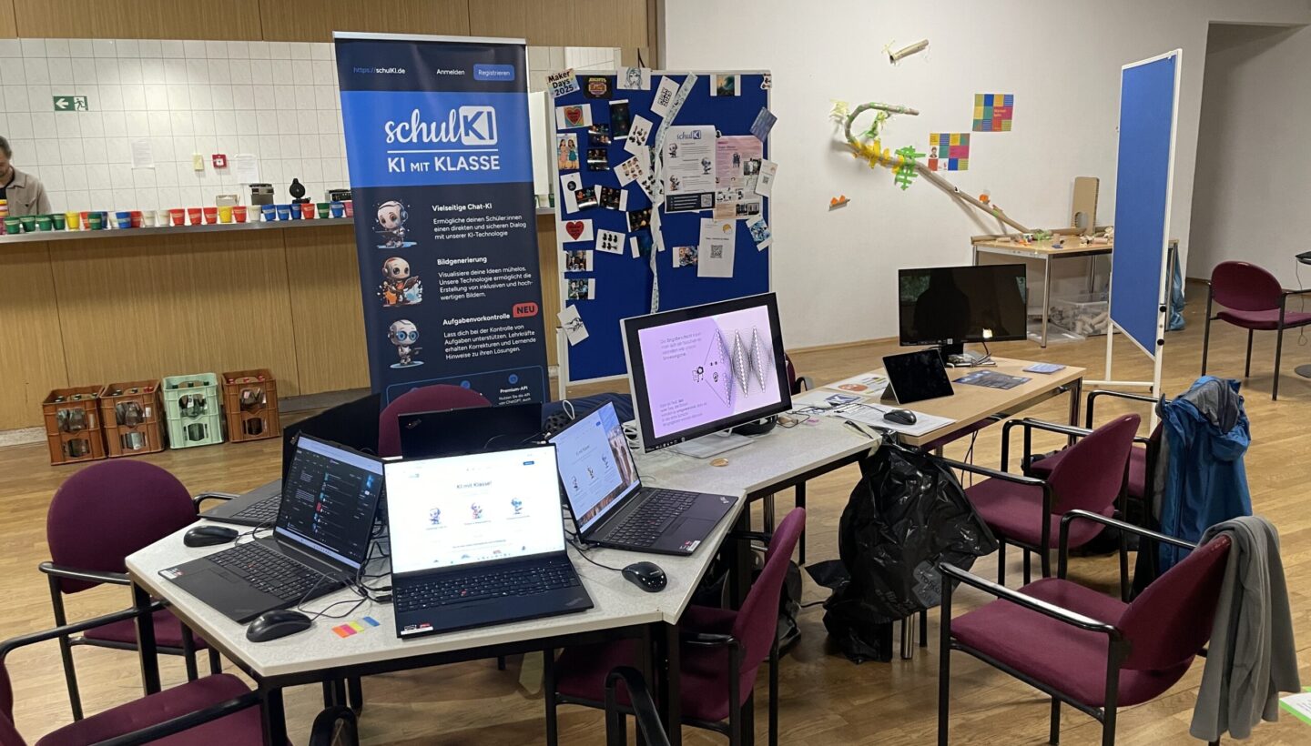 A classroom-style workshop setup with several laptops on tables, a large monitor, and educational materials arranged for participants. A banner in the background reads “schulKI – KI mit KLASSE” with cartoon robots, promoting AI education in schools. A blue display board is covered with papers and notes, and on the right side there is a creative STEM installation with a track or chute. The space is organized and welcoming, designed for interactive learning about artificial intelligence.