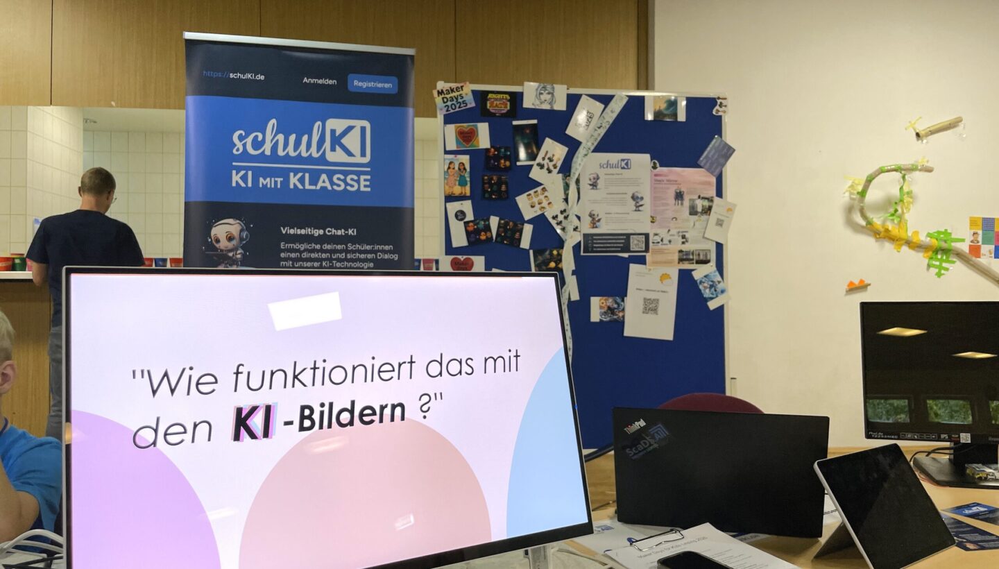 The KI workshop station at the maker days for kids. On a table, a screen displaying the first slide of the slideshow "Wie funktioniert das mit den KI-Bildern?" and notebooks, a tablet and smartphone next to it. Several cluttered items like stamps, a clipboard and flyers are distributed on the table. Behind, a rolltop of schulKI with text; "KI mit Klasse. Vielseitige Chat-KI. ermögliche deinen Schüler:innen einen direkten und sicheren Dialog mit unserer KI-technologie". Next to the rolltop stands a blue partition wall with printed outputs of the KI workshop.
