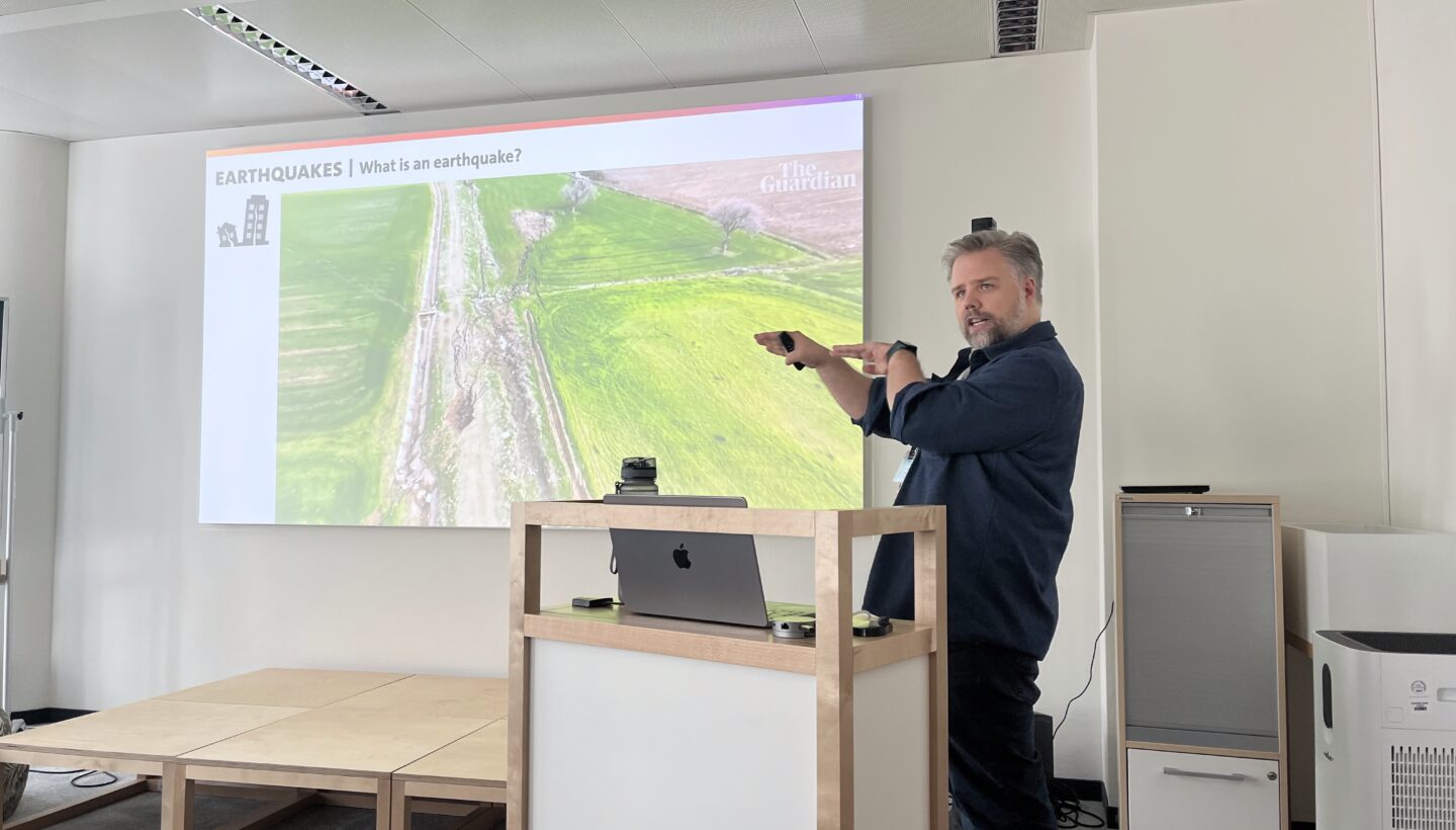 A man gives a presentation on earthquakes, pointing to a projection of a landscape with a visible fault line, in a modern conference room.