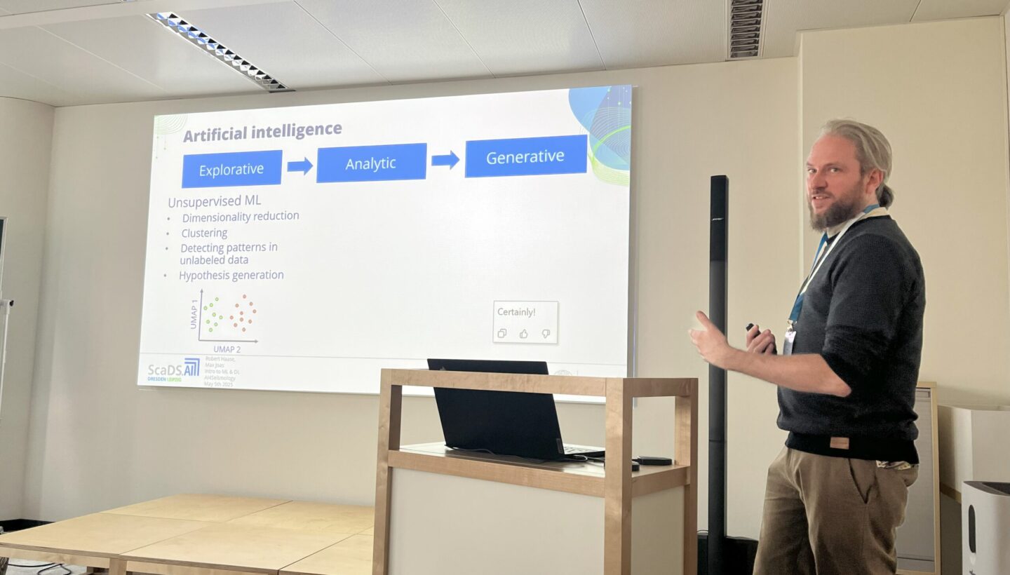 Dr. Robert Haase standing next to a wooden podium. In the background is a presentation projected onto a white wall. The presentation shows a slide about Artificial Intelligence. Three blue boxes connected with arrows, within is one word each: "Explorative", "Analytic", "Generative". Below is a headline: "Unsupervised ML" and several bullet points: "Dimensionality reduction", "Clustering", "Detecting patterns in unlabeled data" and "Hypothesis Generation". Lastly, there is a graph with two axes. The x axis is labeled "UMAP 2" and the y axis "UMAP 1". Data points in green are clustered around the lower UMAP 2 value and all the way up on UMAP 1, next to this are red data points clustered on the higher scale uf UMAP 2 and also all the way up on UMAP 1. The green and red data points are clearly seperated.