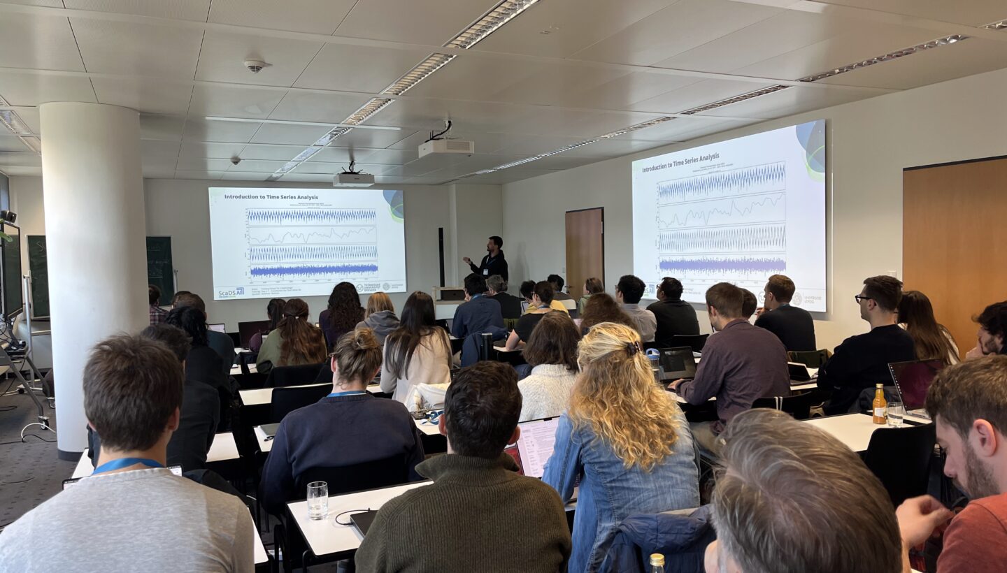 A classroom full of people. The perspective shows the people from behind and Matthias Täschner holding a presentation in the back. The slides projected on the white wall shows "Introduction to Time Series Analysis" and 4 different seismic records.