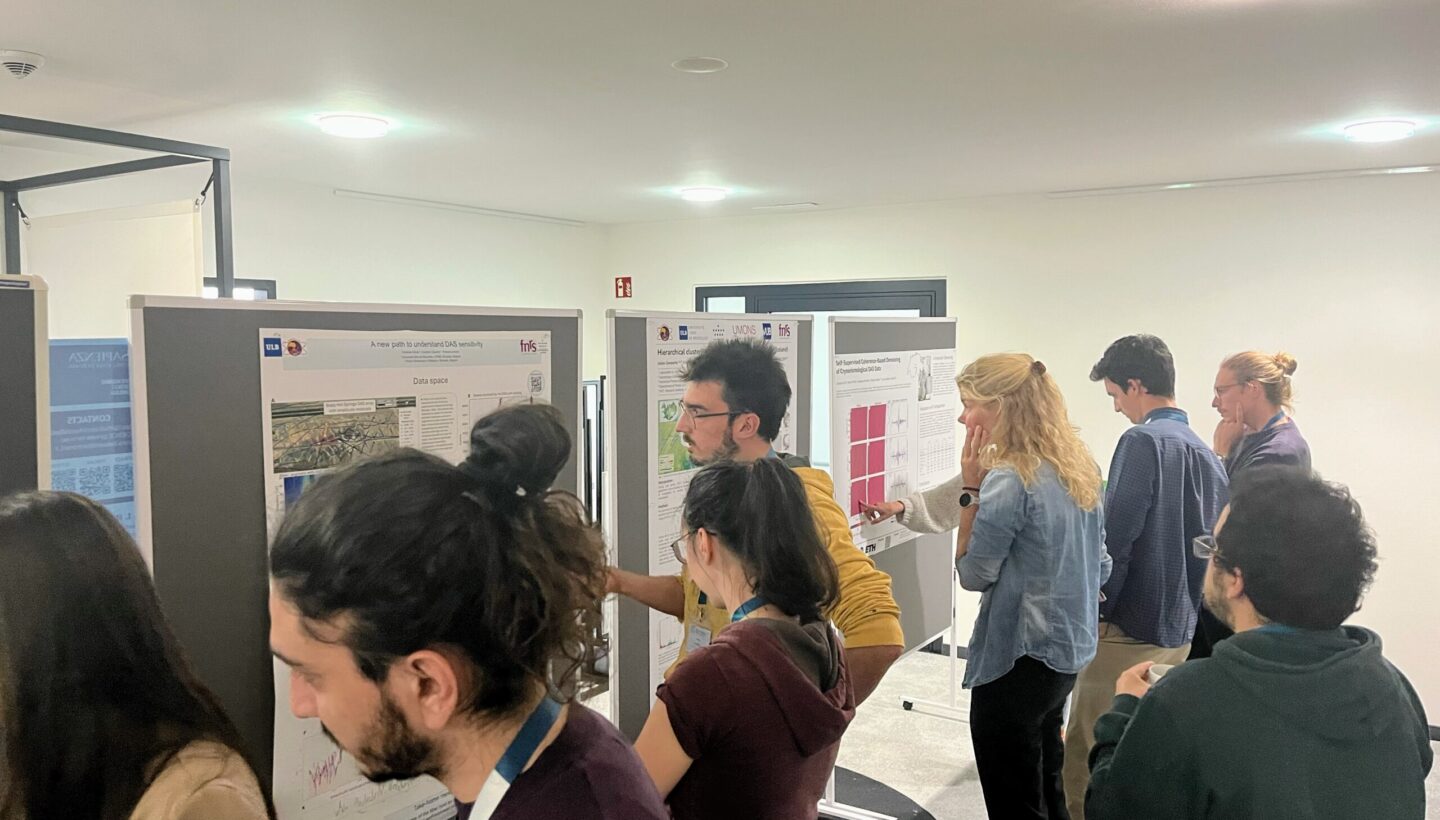 A poster session during the AI4Seismology Training. Inside a white room, diagonally placed are three grey partition walls with posters on them. Before and spread out through the room are several people. All looking concentrated at the posters.