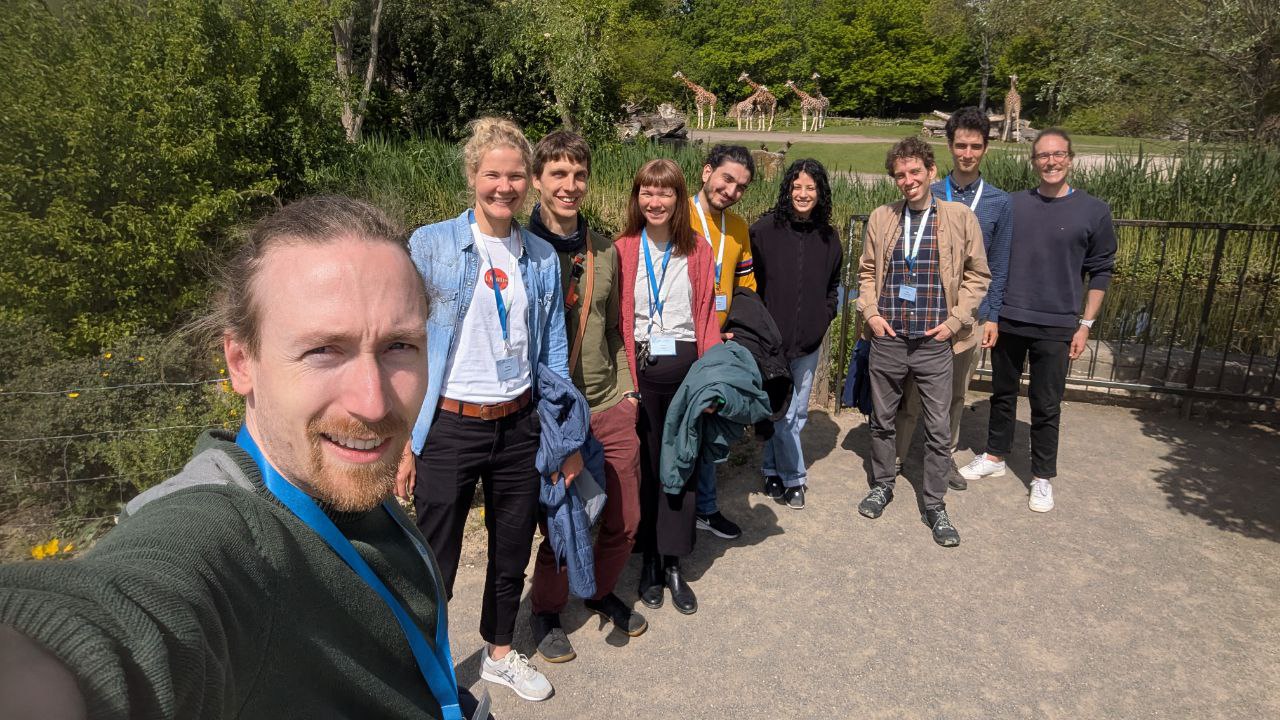 A selfie with nine people. They are all smiling into the camera. In the background the Leipzig Zoo. A lot of green and some giraffes.