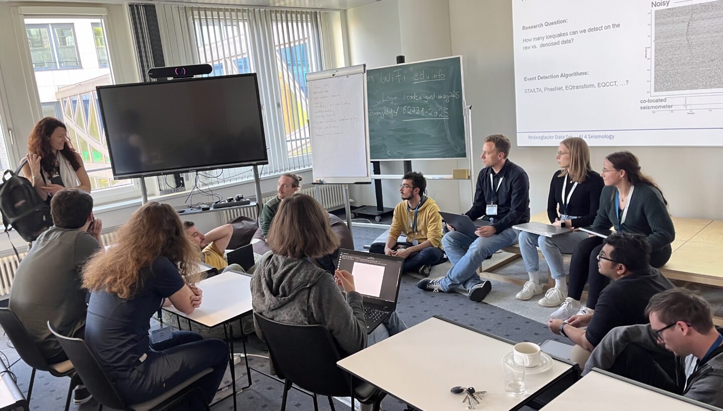 A group of twelve people gathered in the living lab Leipzig. Sitting on the floor, on charis or on a small wooden stage. Most of them have a notebook in front of them. They are casually clothed. The picture shows the left corner of the room. There is a presenter TV and a chalkboard, behind those is a windowfront.