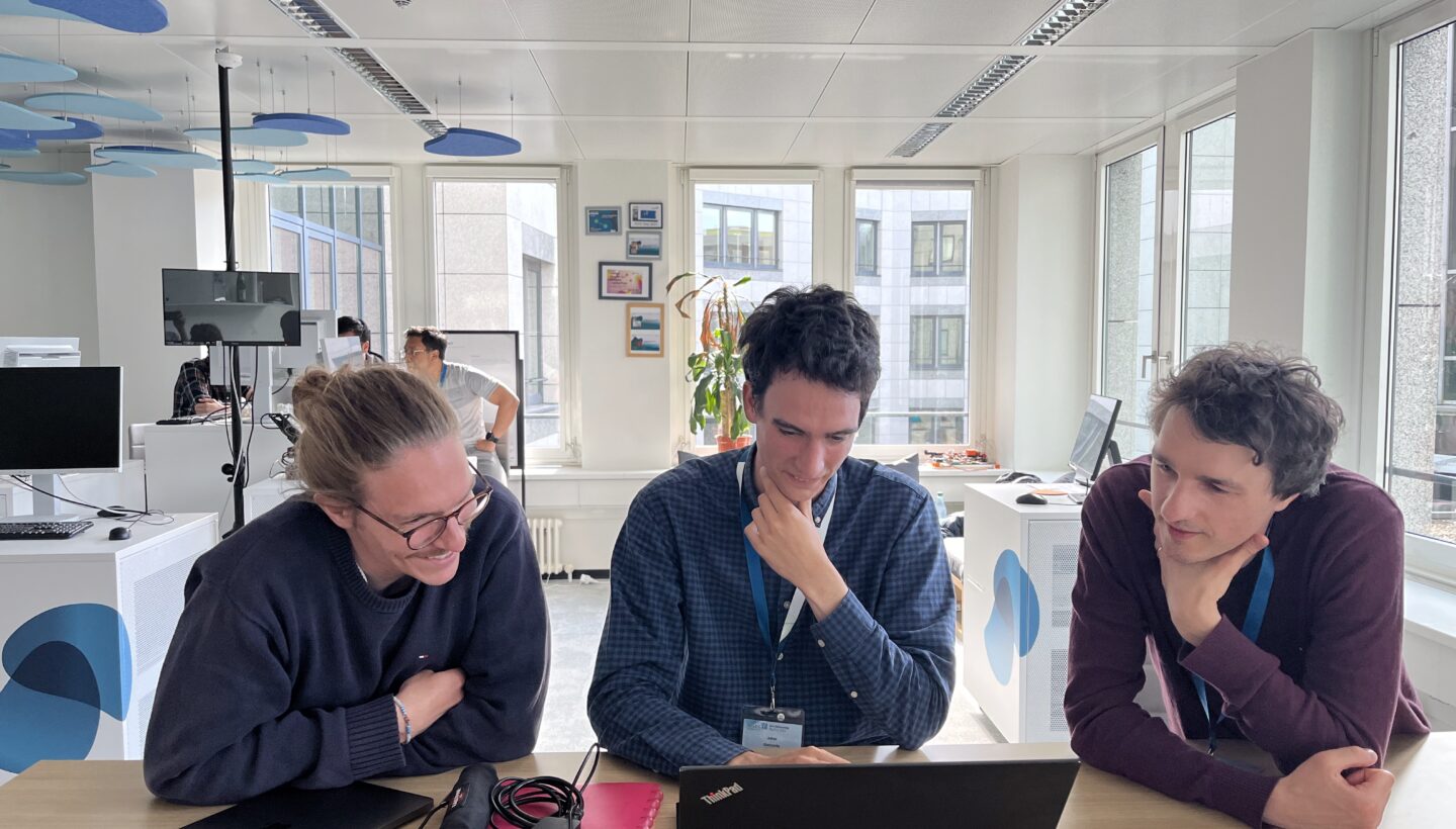 In the living lab leipzig. Three people looking sitting in front of a wooden table, looking at a notebook. The person on the left smiles the other to seem very interested and engaged in what is shown on screen.
