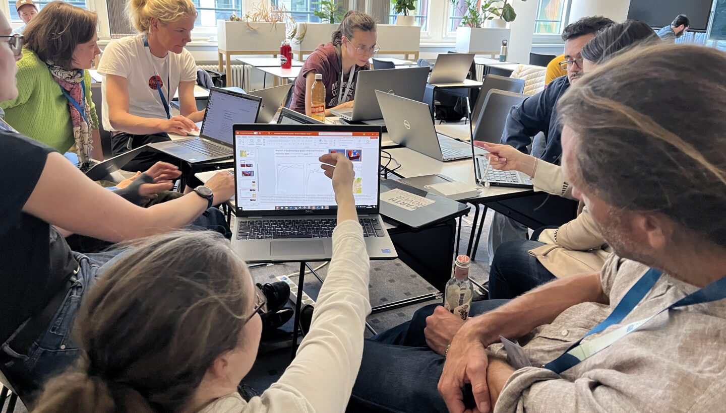 A group of people sitting closely gathered around a few small tables with notebooks on them. 2-3 people look at one notebook and one person explains the data shown on the notebook screen.
