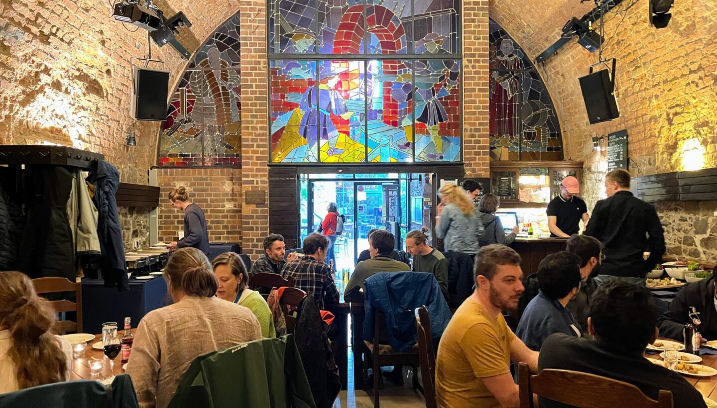 Inside the Moritzbastei Leipzig. The walls and ceiling are built of bricks and form a half circle. In the back, there is colorful big glass window and a glass door below. Distributed through the room on tables are people eating and talking to each other.