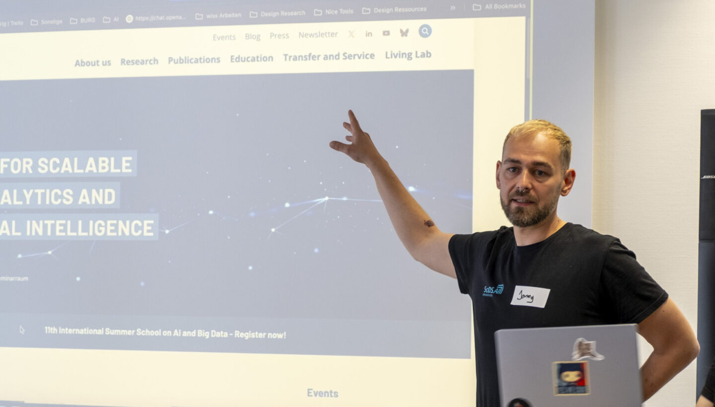 Johannes Häfner stands at a podium presenting the ScaDS.AI homepage on a large projection screen. The screen displays the website's navigation and content related to technology transfer, education, and upcoming events.