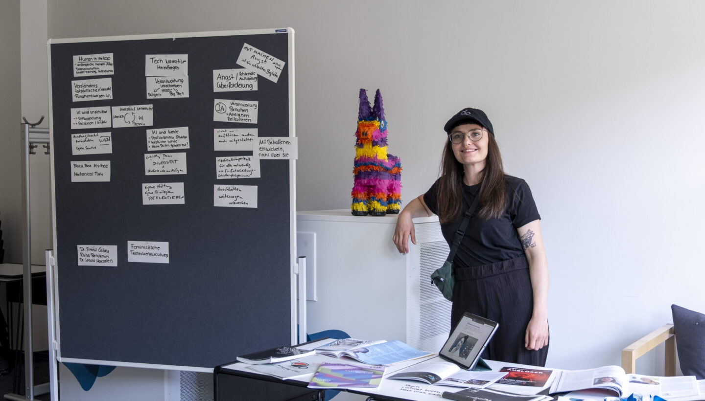 Vanessa Kuhfs stands in front of a literature table featuring books and magazines referenced during her talk. Adjacent to the table, a board displays the group work results with participant responses summarized by academic discipline.