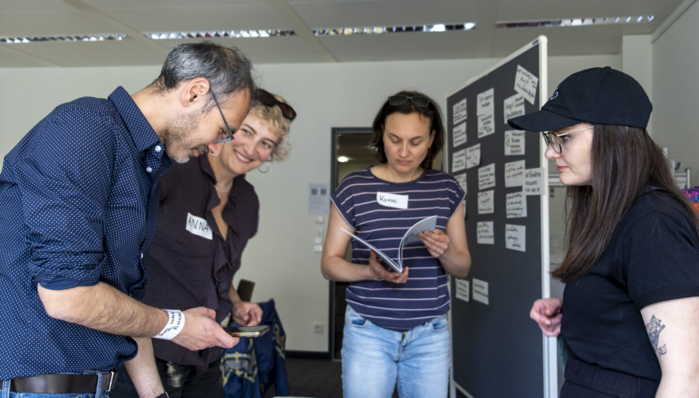 Participants actively browse a literature table displaying an array of books and magazines referenced in Vanessa Kuhfs’ presentation. The table is neatly arranged with academic texts focused on artificial intelligence and ethics, and several attendees are seen examining the materials in a well-lit conference space.