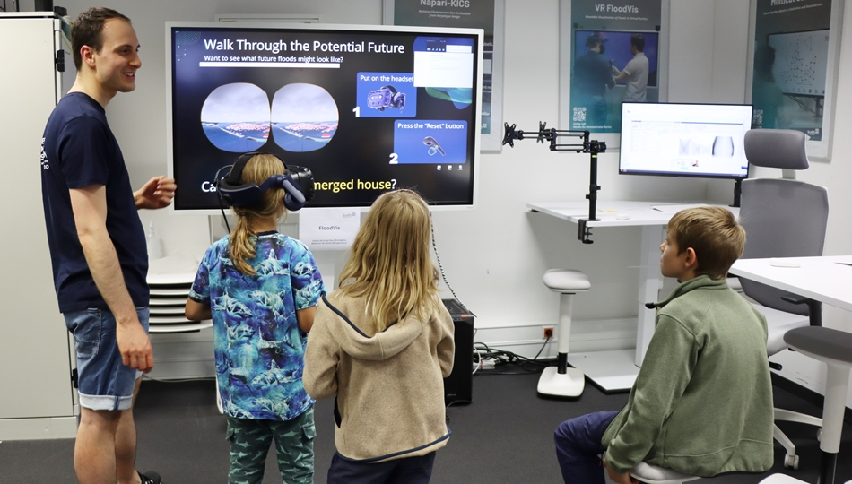 Photo. Moritz Müller is explaining the FloodVis VR demo to three children at the Living Lab in Dresden.