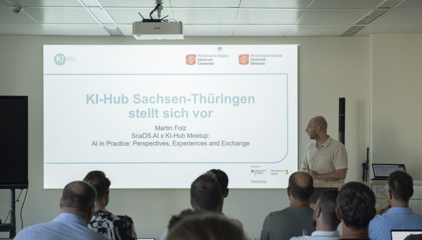 At a ScaDS.AI KI-Hub Meetup, Martin Folz speaks in front of a slide titled “KI-Hub Sachsen-Thüringen stellt sich vor” with the subtitle “AI in Practice: Perspectives, Experiences and Exchange,” flanked by logos of ScaDS.AI Dresden/Leipzig, Mitteldeutscher Digital Zentrum Chemnitz, and Ilmenau, as attendees listen.