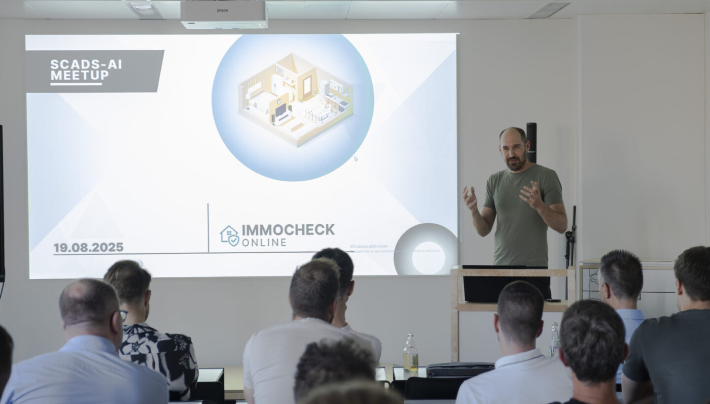 Speaker at the SCADS-AI Meetup stands beside a podium gesturing to a projected slide titled “IMMOCHECK ONLINE” dated 19.08.2025, which displays a circular graphic of a stylized house interior; an attentive audience is seated in a modern conference room.