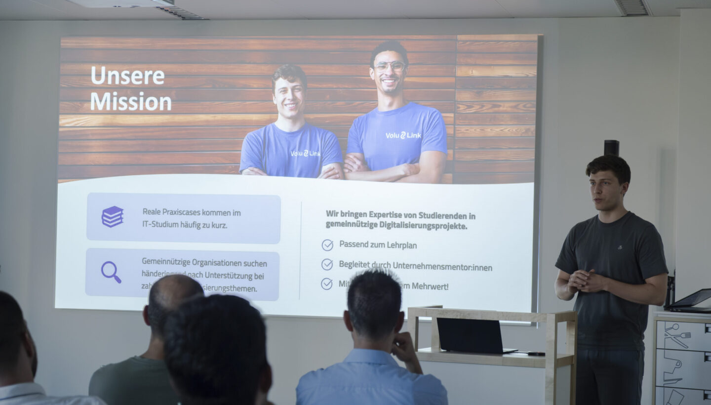 Presenter stands next to a wooden podium addressing attendees, with a slide titled “Unsere Mission” behind him showing two smiling people in blue Volu Link shirts and bullet points describing the integration of student IT expertise into nonprofit digitalization projects.