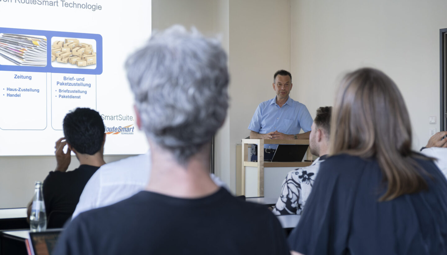 Presenter behind a podium speaks to an audience as a slide titled “RouteSmart Technologie” shows two sections: left, an image of newspapers labeled “Zeitung” with house delivery and trade; right, parcels labeled “Brief- und Paketzustellung” with letter and parcel details, and SmartSuite and RouteSmart logos at the bottom.