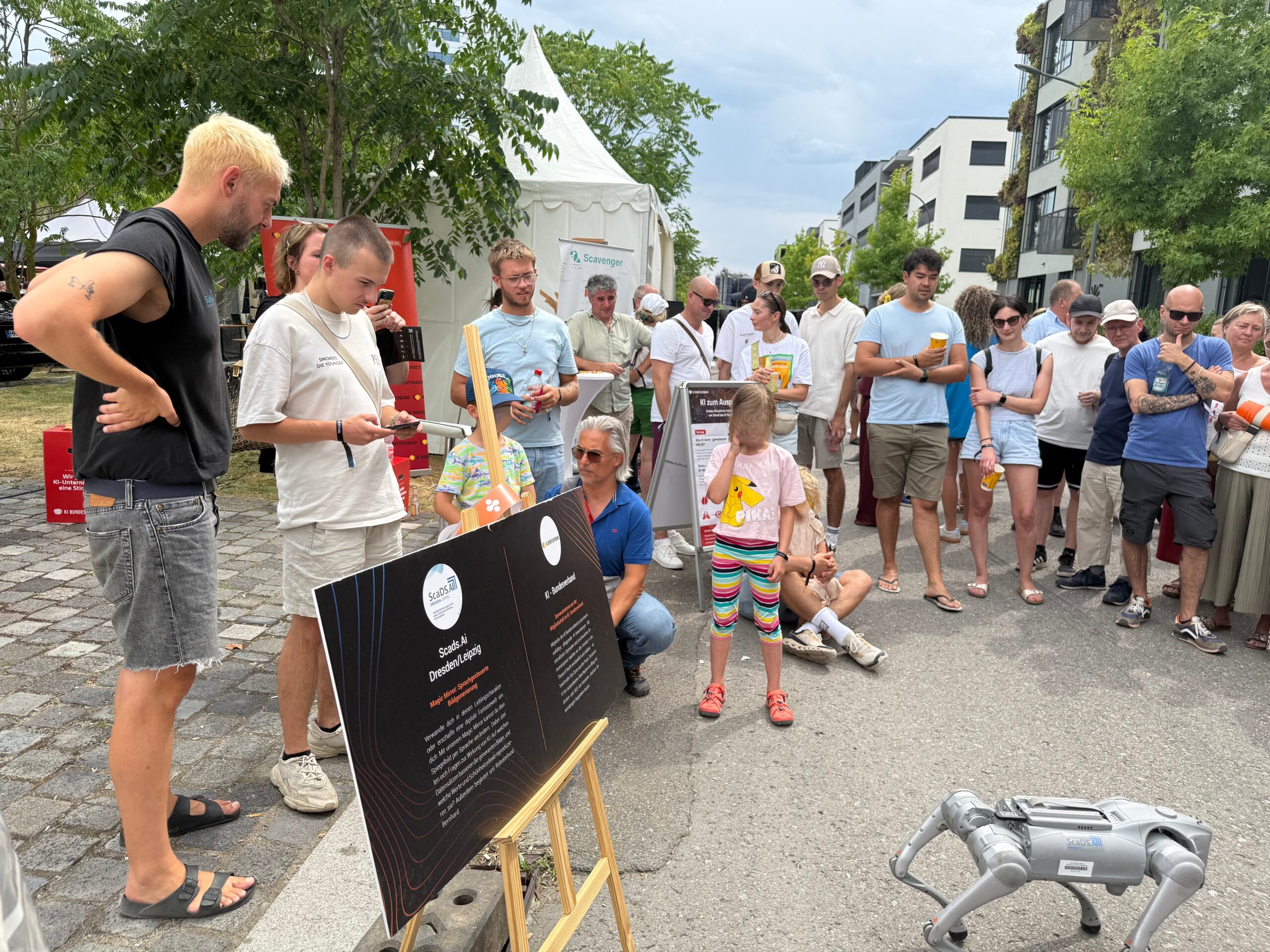 A group of people standing outside next to a white tent. They all look in direction of the ground where a Quadro cop is standing. In the background are some trees and houses. To the left there is one person controlling the Quadro cop.