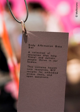 Photo. Tag with inscription:

"Tag with Body Affirmation Data Set.

A collection of resources that help Trans* and autistic people thrive in our bodies.

This contains health care resources, DIY recipes for emodied access tools, and open questions."

© Meltionary