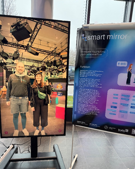 Photo. Johannes Häfner and Vanessa Kuhfs take a photo of their reflection in the Magic Mirror. Next to the display, there is a poster about the demonstrator.