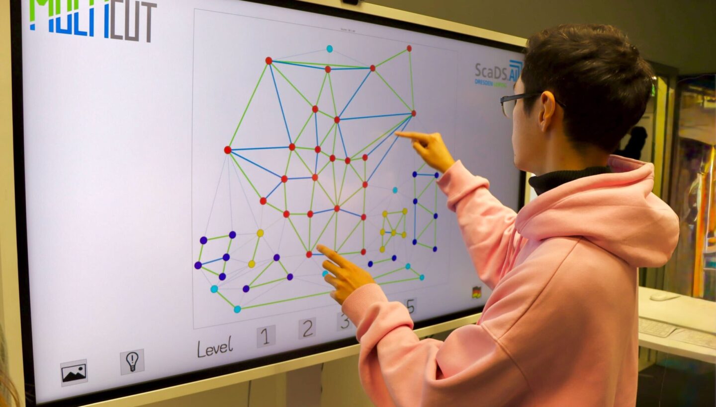 Photo. A guy in a pink hoodie stands at a large monitor, using his fingers to trace and point at a diagram of colorful dots and lines,  solving a Multicut algorithms.