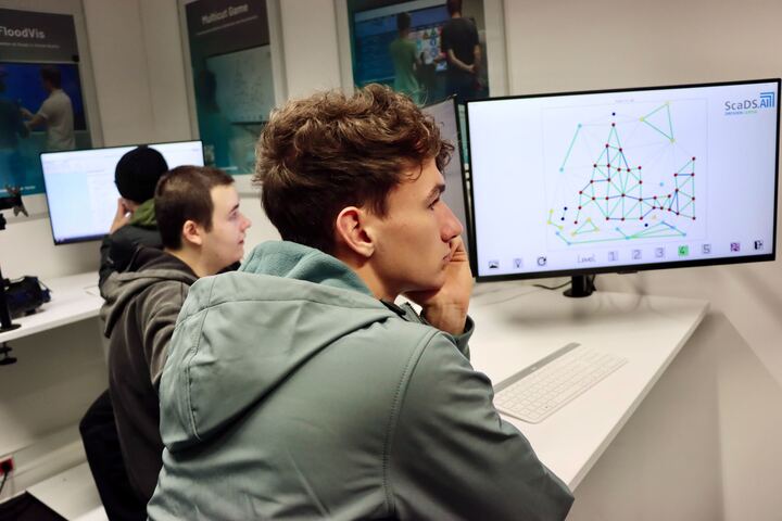 Photo. A guy in a grey hoodie rests his chin on his hand, concentrating on a large monitor that displays a graph. Another guy sits next to them, also looking at a screen in a Living lab.