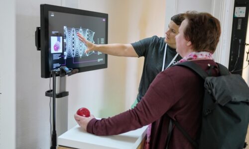 Photo. Monika Reich is presenting the asanAI demonstrator to a female visitor of the Building Bridges 2025 conference. The woman holds an apple in her hand, which was used to train data in the asanAI demo.